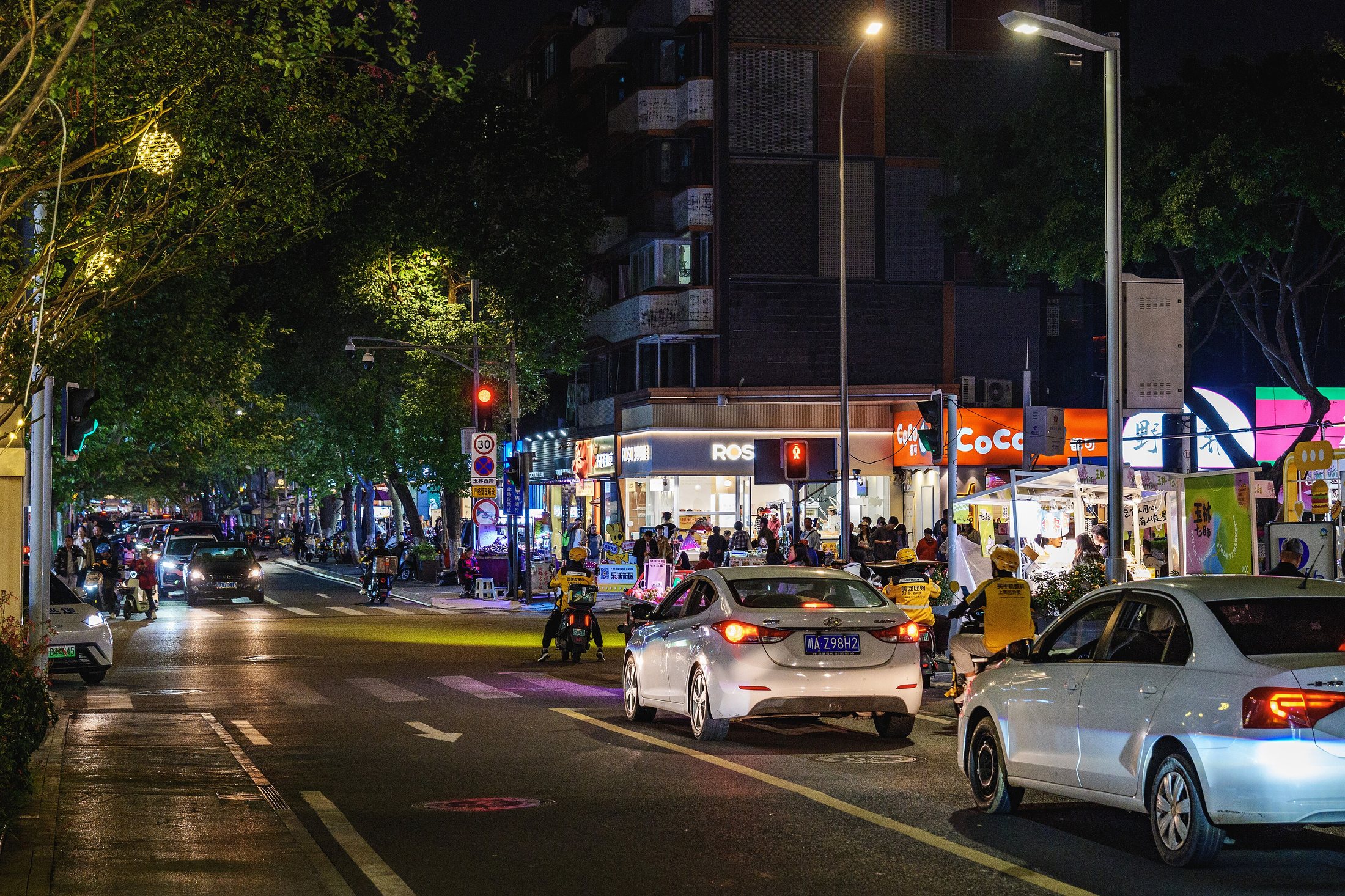 A lively street intersection in Chengdu's Yulin area with trees, shops, and evening city life.