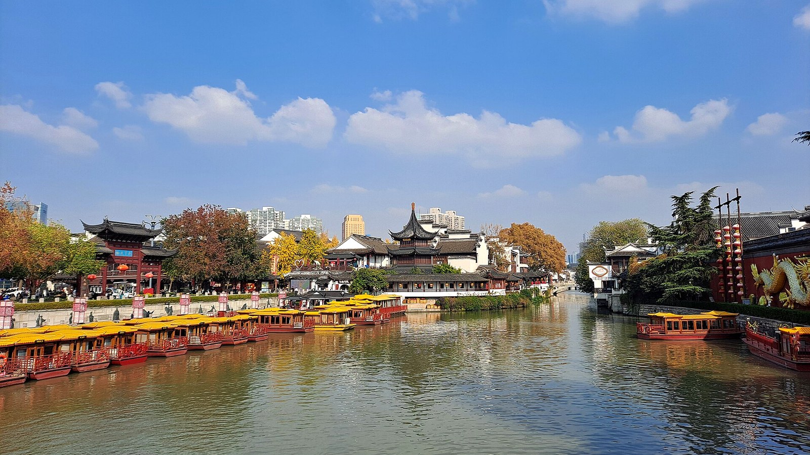 Daytime view of the Qinhuai River in Nanjing with traditional riverside buildings and sightseeing boats under a bright blue sky.