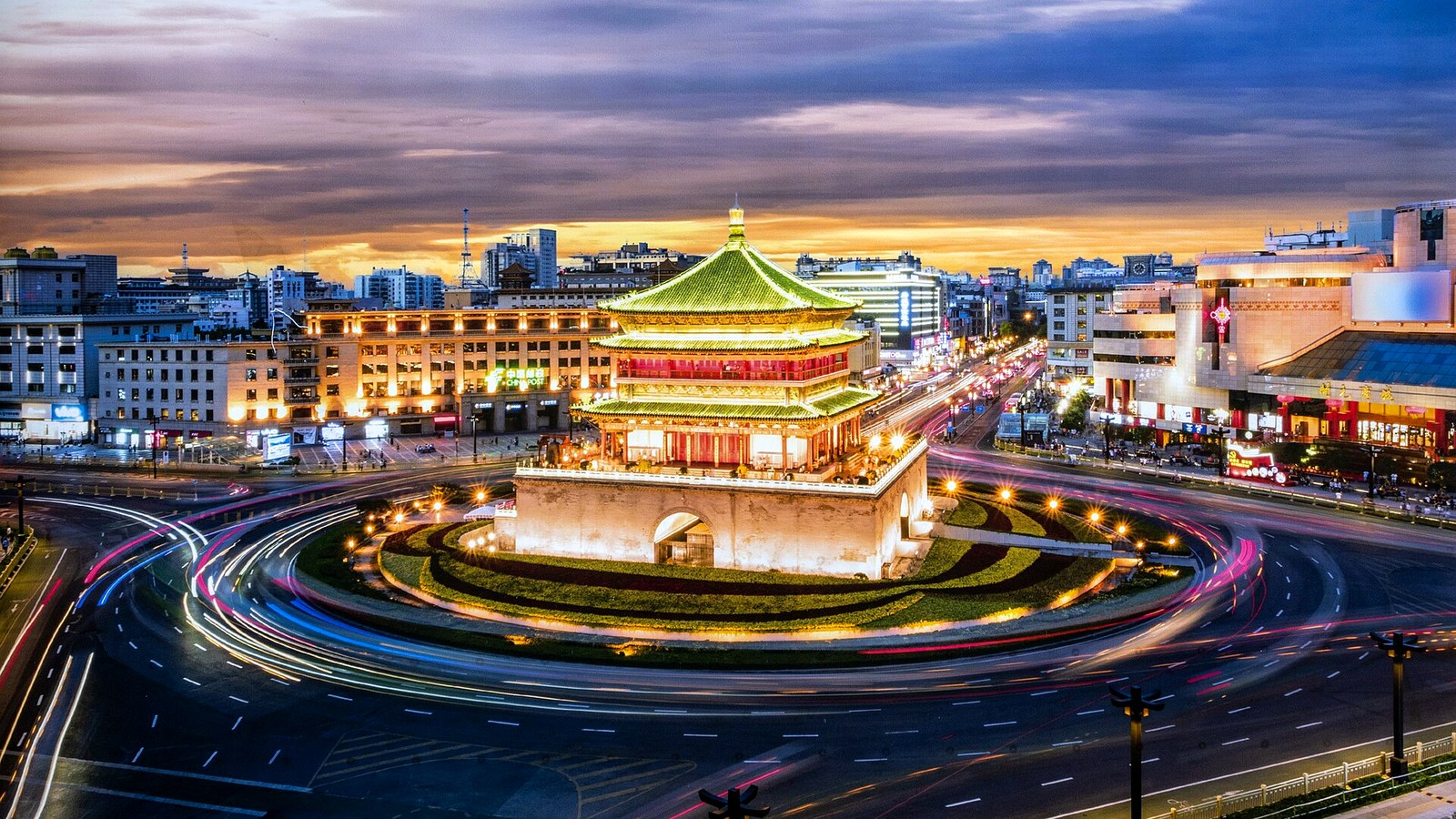 Xi'an Bell Tower glowing at dusk with sweeping traffic light trails circling the roundabout beneath a dramatic sky.