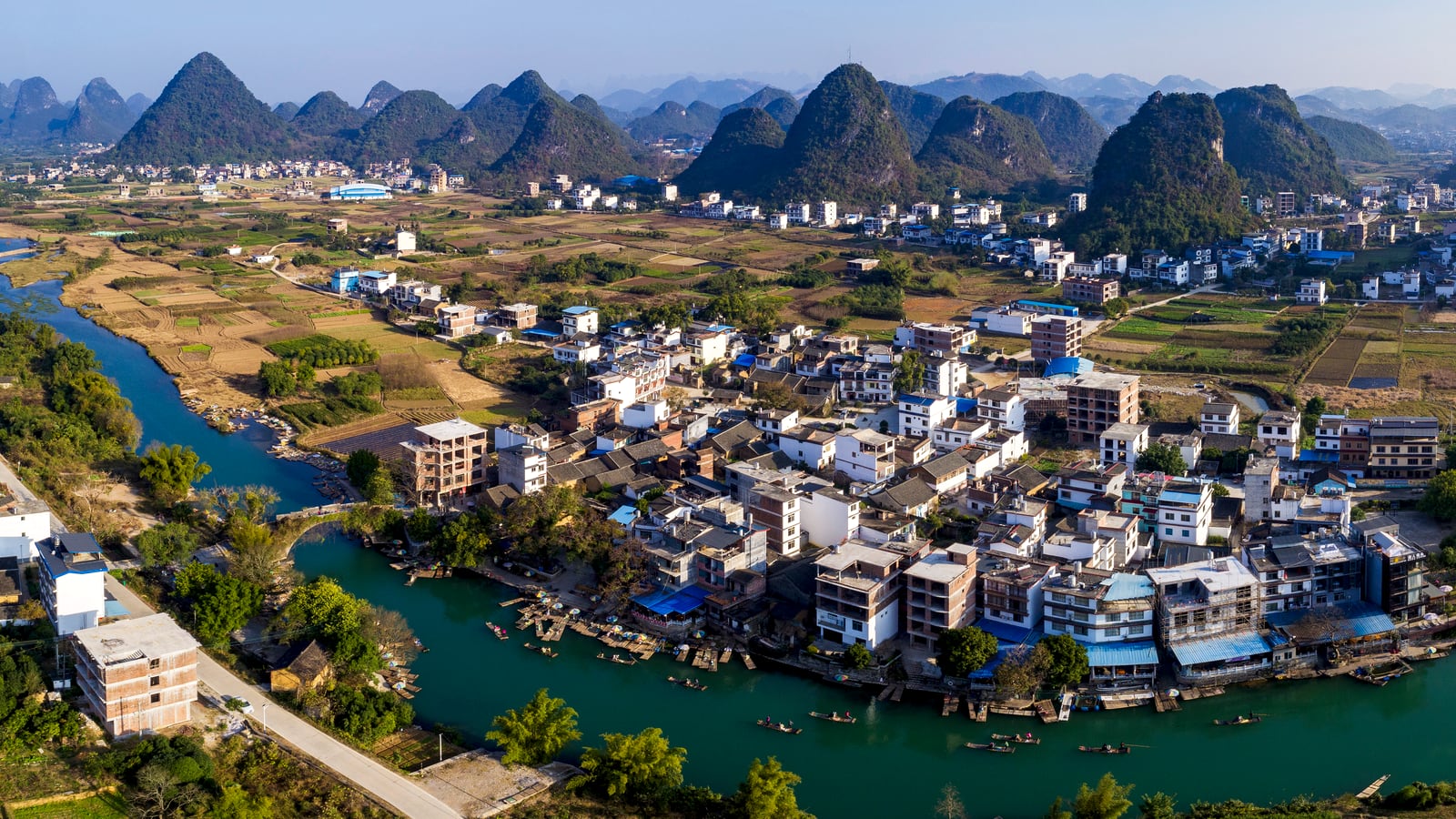 A bright aerial view of a river bend, village rooftops, and karst peaks around Yangshuo.