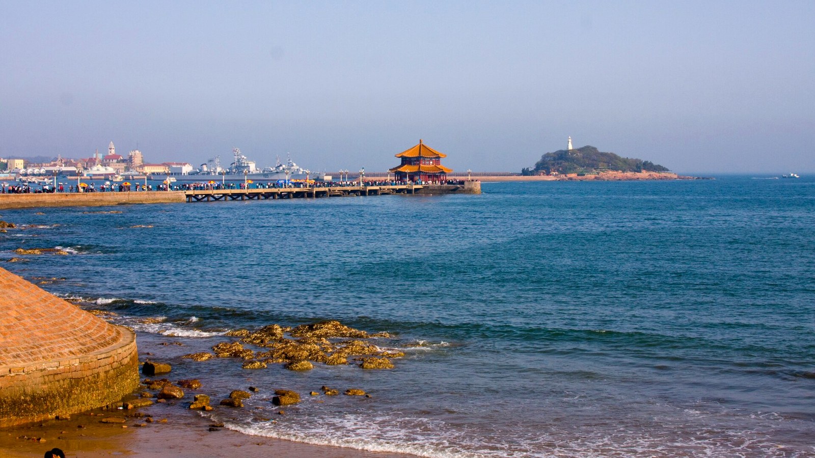 Zhanqiao Pier in Qingdao reaching toward Huilan Pavilion, with blue water and Little Qingdao beyond.