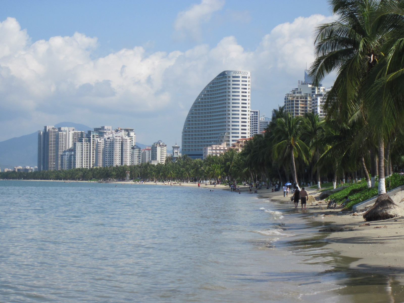 Sanya Bay under clear light with curving shoreline, palms, and high-rise resort buildings behind the beach.